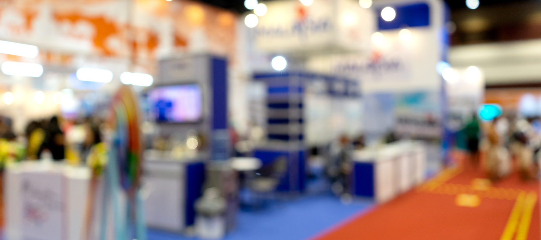 Blurred image of a trade show with booths, colorful displays, and bright lighting. People are present, creating a bustling and energetic atmosphere.